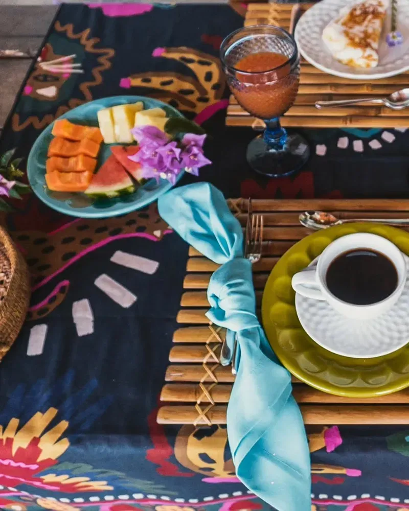 Uma mesa de café da manhã com uma toalha estampada vibrante, com frutas, café, suco e jogos americanos sobre esteiras de bambu.