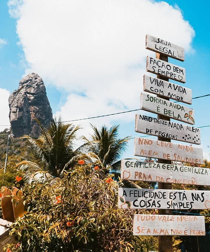 Um poste vertical com várias placas de madeira contendo mensagens positivas, tendo como pano de fundo um pico rochoso e um céu azul.