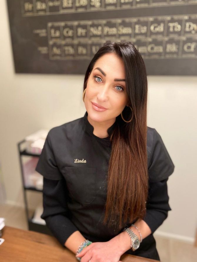 Una donna dai lunghi capelli scuri, con indosso una maglietta nera, è seduta al chiuso e sorride in una sala per trattamenti.