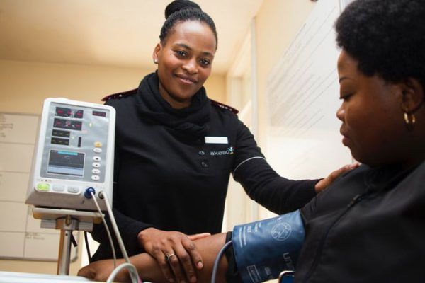 A healthcare worker in a dark uniform monitors a patient's blood pressure using a digital machine in a clinical setting.