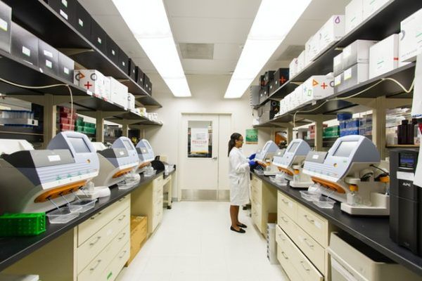 A researcher in a lab coat works at a station with several diagnostic devices lining the counters of a brightly lit lab.