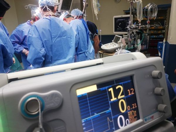 Medical staff in scrubs perform surgery in an operating room, viewed from behind a patient vital signs monitor.