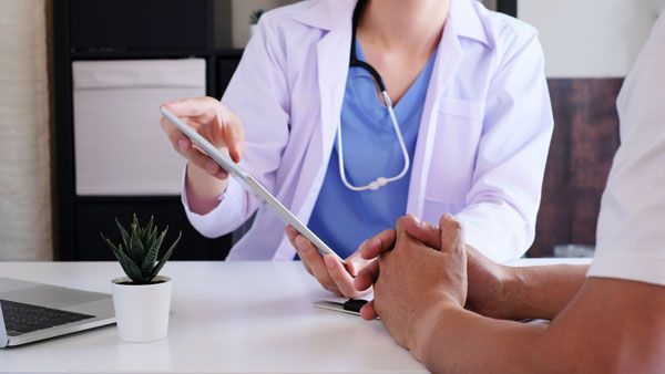 A medical professional in a white coat and blue scrubs uses a tablet to explain information to a patient in an office.