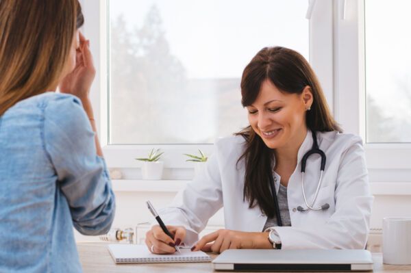 A doctor in a white coat with a stethoscope around her neck writes notes while consulting with a patient in an office.