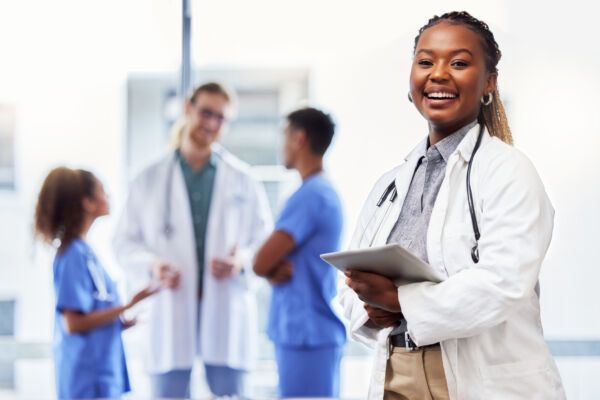 A smiling doctor in a white coat holds a tablet, with blurred healthcare colleagues talking in the background.