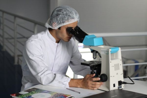 A person in a white lab coat and hairnet looking through a microscope in a professional setting.