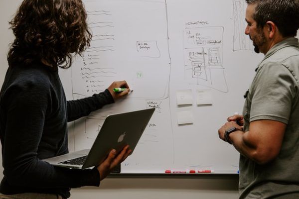 Two people collaborate at a whiteboard; one writes with a marker while holding a laptop, the other observes the board.