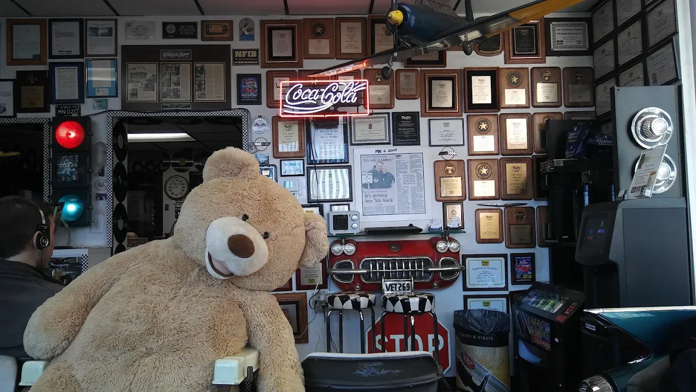 Teddy bear in a shop, in front of a wall covered in framed awards, with a red/green traffic light and Coca-Cola sign.