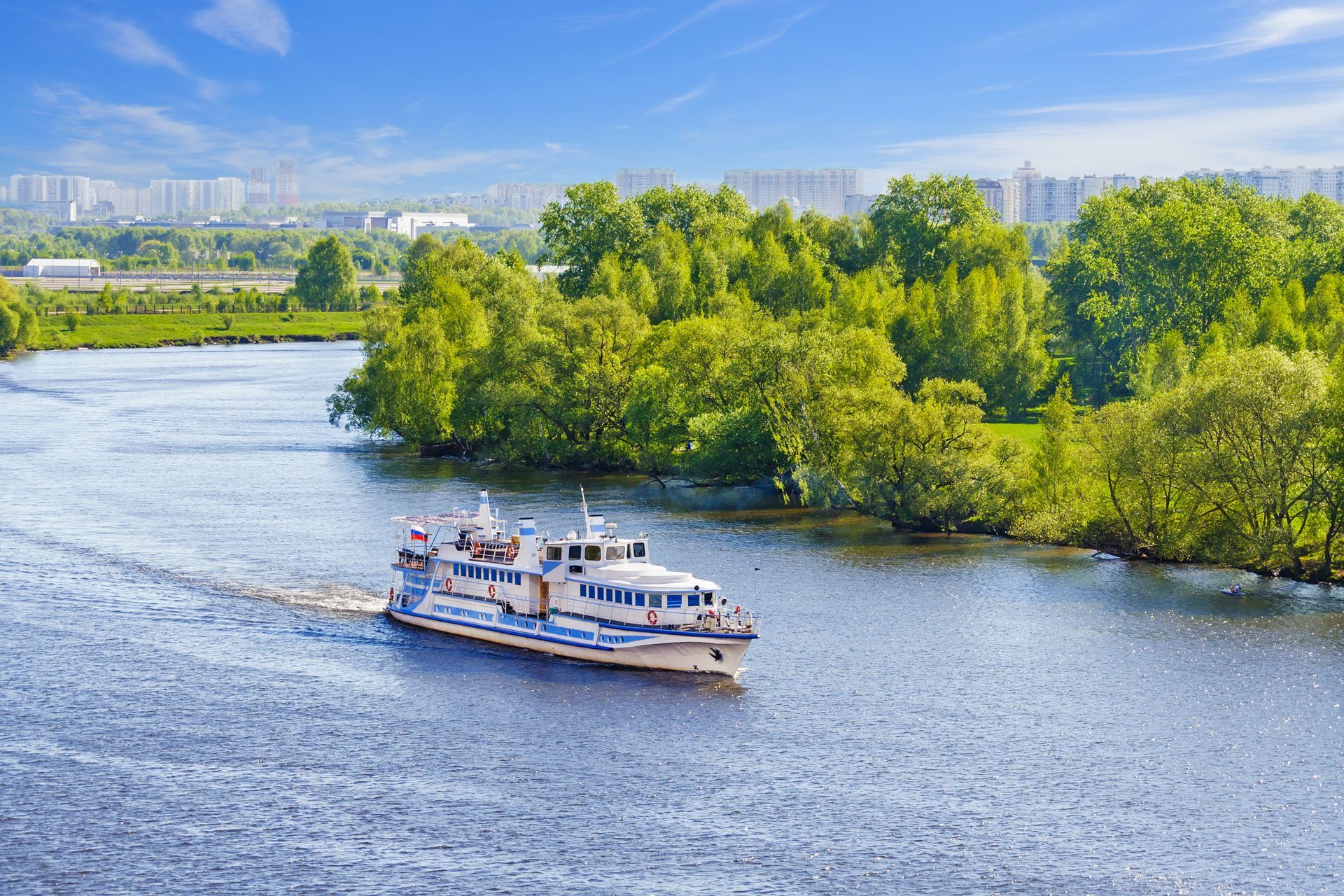 Boat traveling on a river, surrounded by lush green trees and a distant cityscape under a blue sky. Boat traveling on a river, surrounded by lush green trees and a distant cityscape under a blue sky.