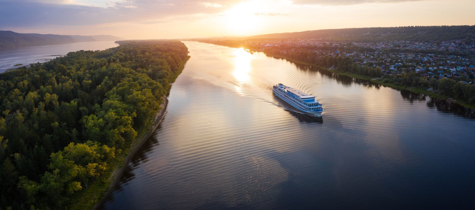 River cruise ship sailing on a wide river at sunset. A lush green island is on the left. River cruise ship sailing on a wide river at sunset. A lush green island is on the left.