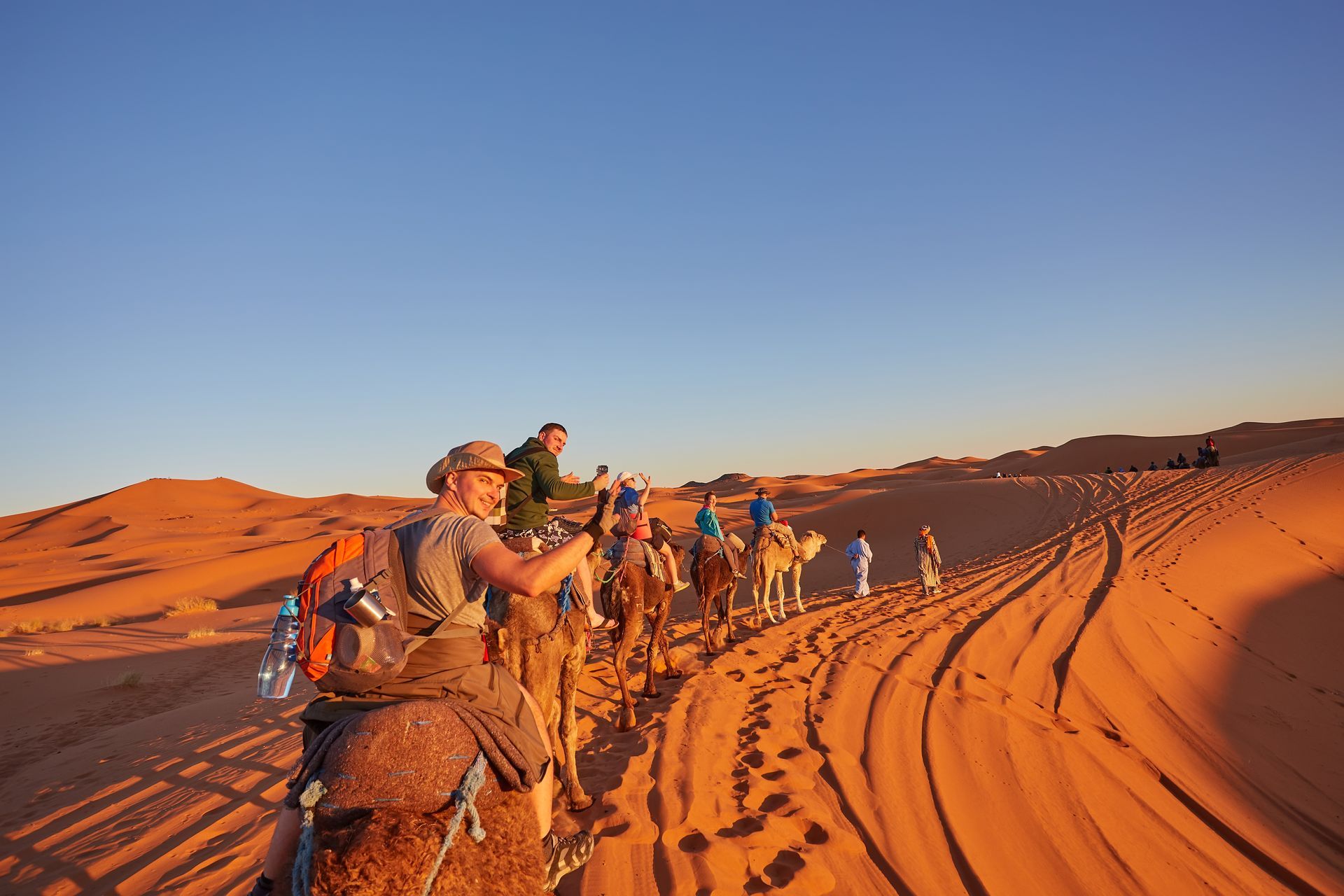 People ride camels across desert dunes under a blue sky, tracks in the sand. People ride camels across desert dunes under a blue sky, tracks in the sand.