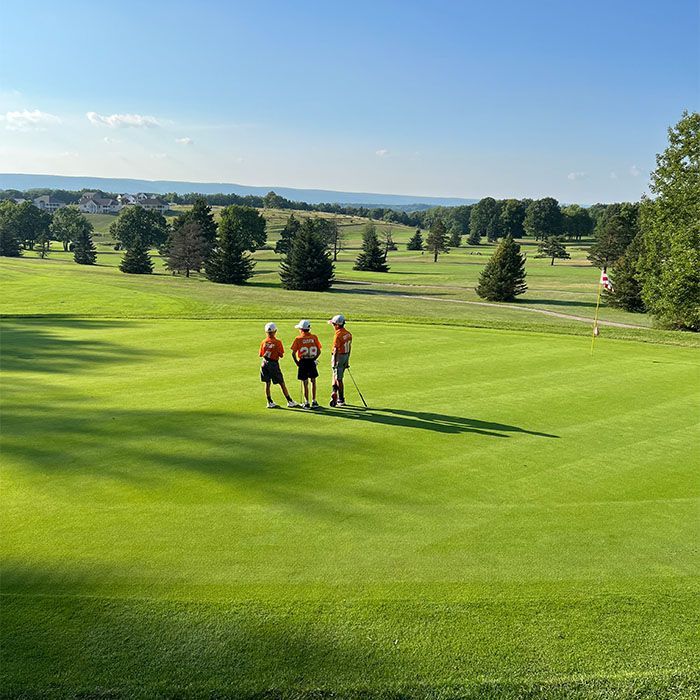 Three golfers in orange vests walk across a sunlit green golf course with trees and hills in the distance