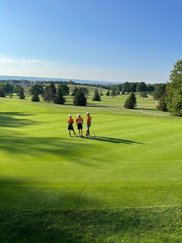 Three people in orange shirts walking across a bright green golf course under a blue sky.