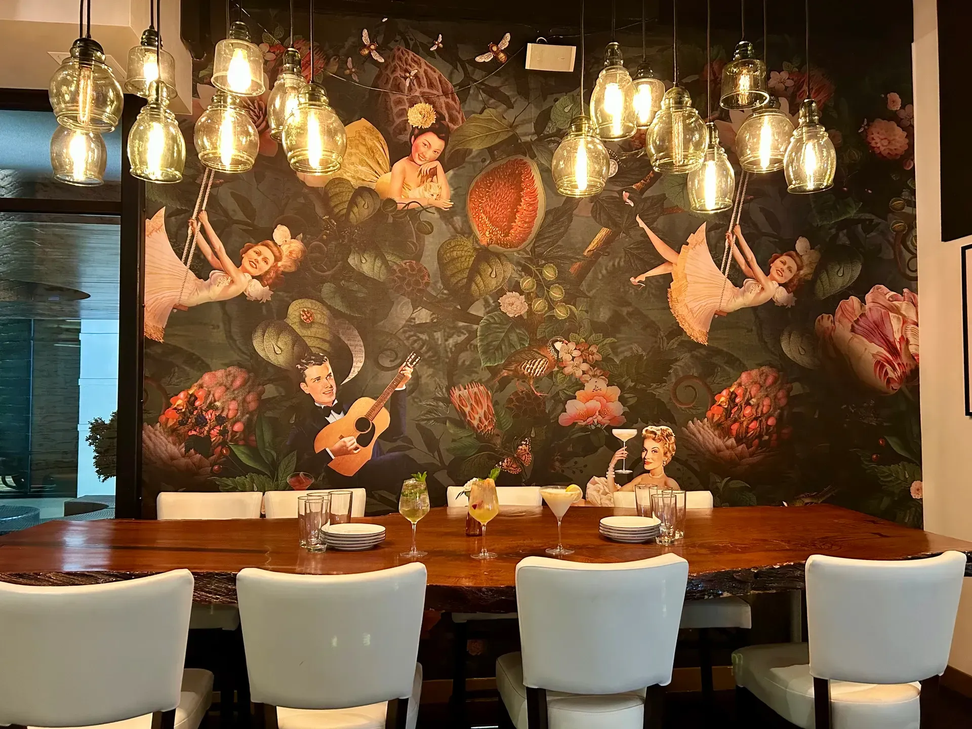 Dining room with large wood table, white chairs, whimsical mural, and hanging lights.
