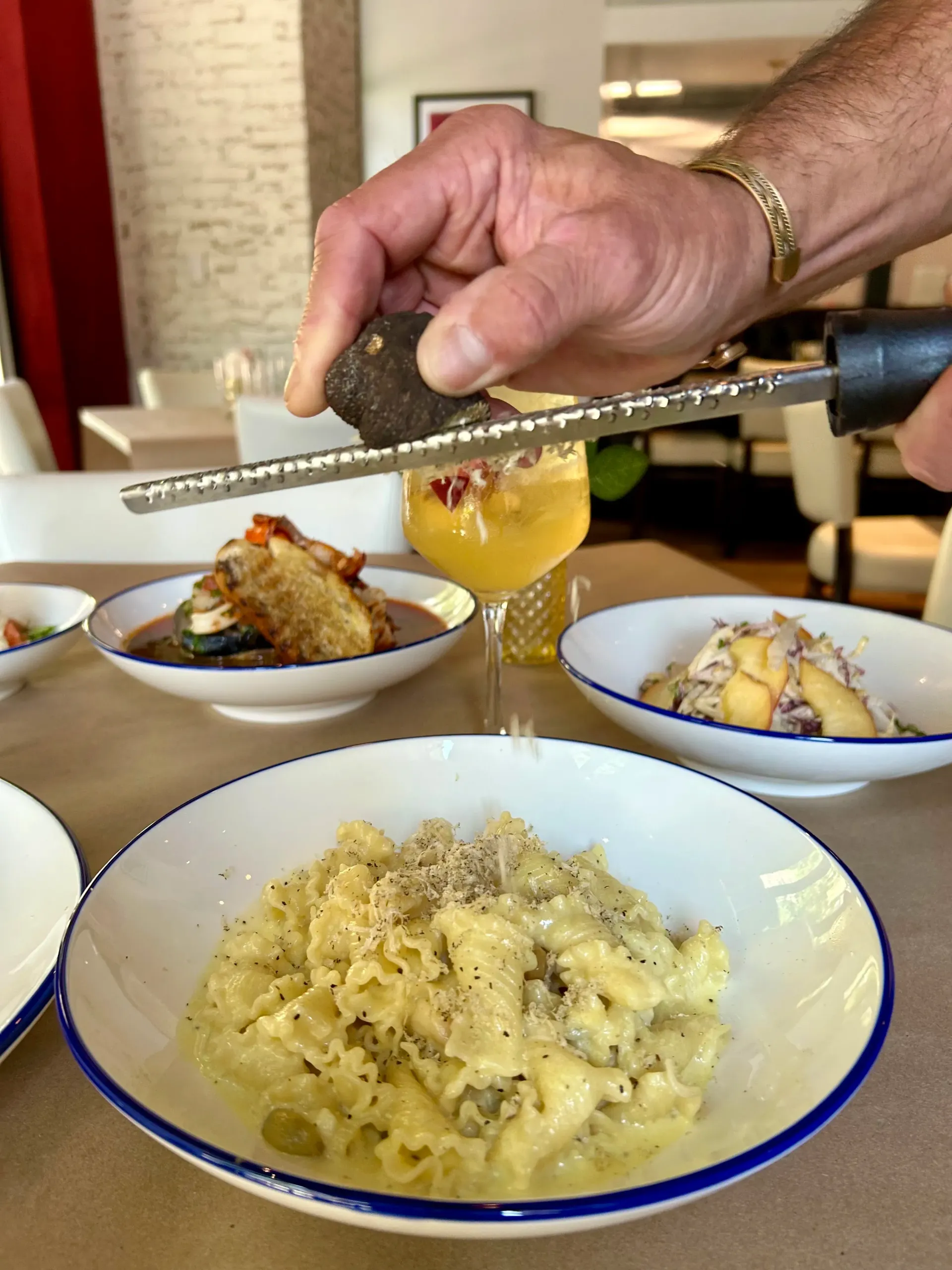 Chef grating truffle over pasta dish at a restaurant table.