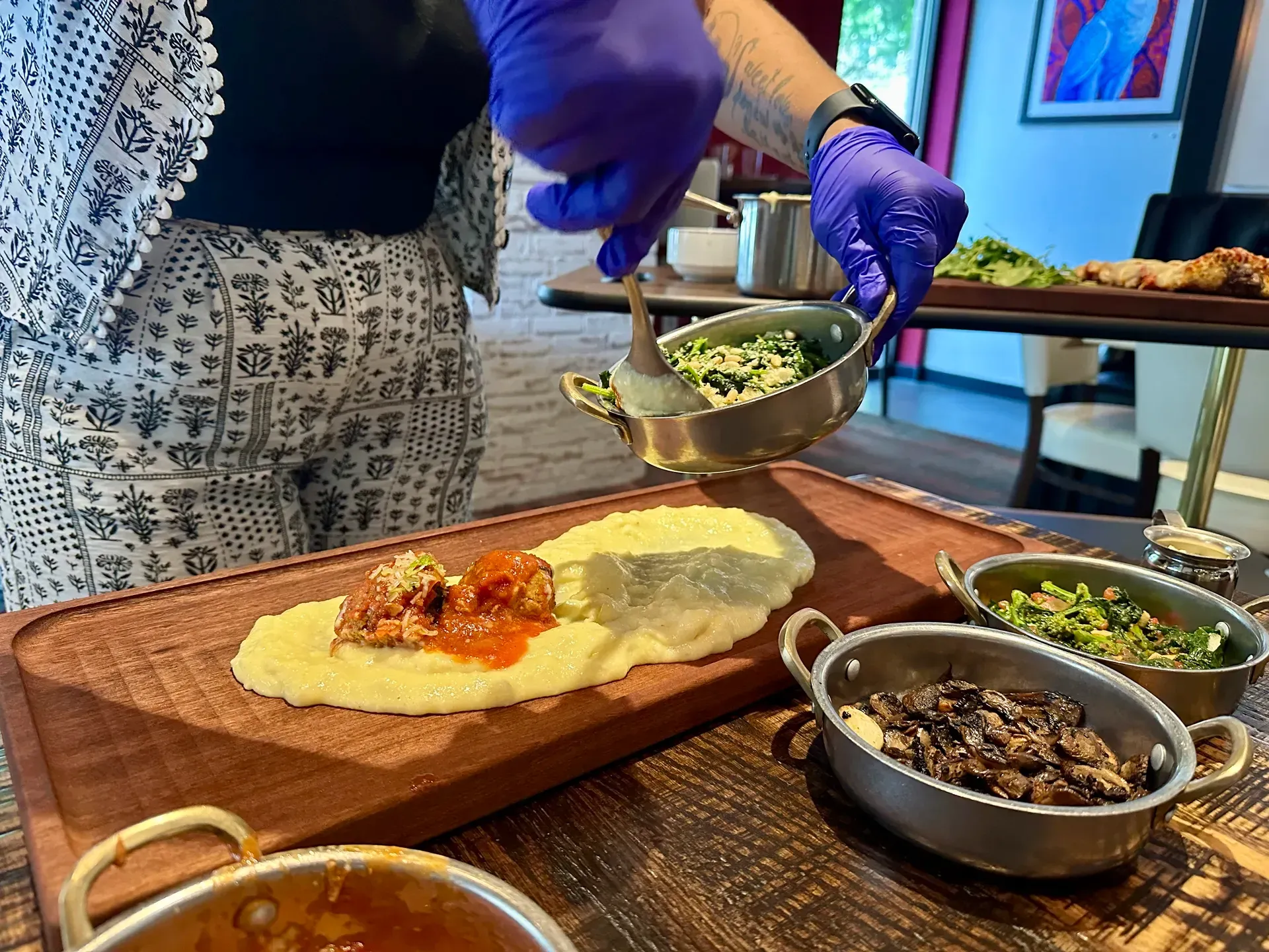 Person with blue gloves plating food onto a wooden board with several side dishes.
