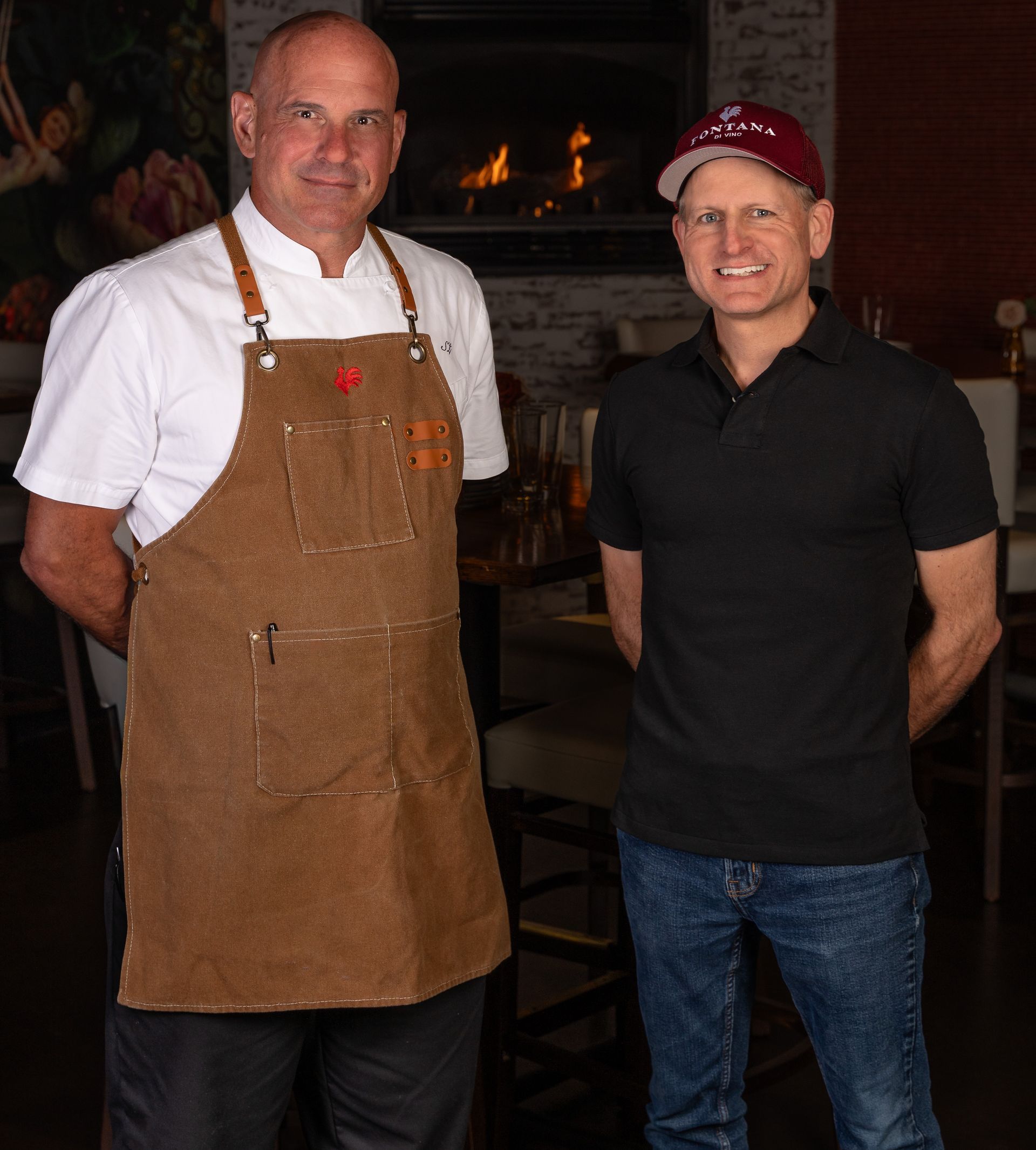Chef and man standing indoors; chef wears apron; fireplace in the background.