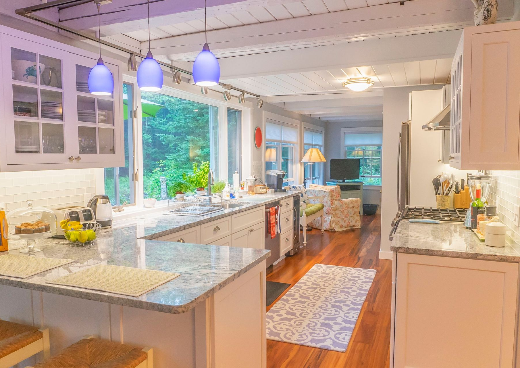 Bright, airy kitchen with white cabinets, granite countertops, blue pendant lights, and a view of lush greenery.