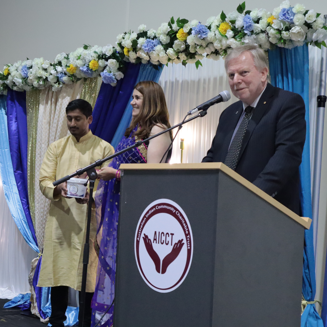Three people on a decorated stage, with a man speaking at a podium under floral drapes.
