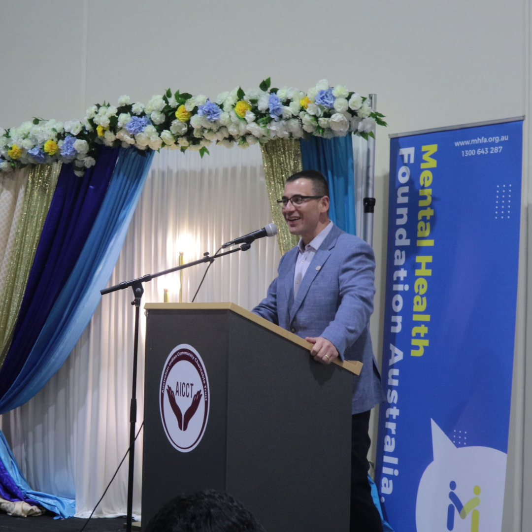 Man speaking at a podium on a decorated stage with a blue Mental Health Federation Australia banner