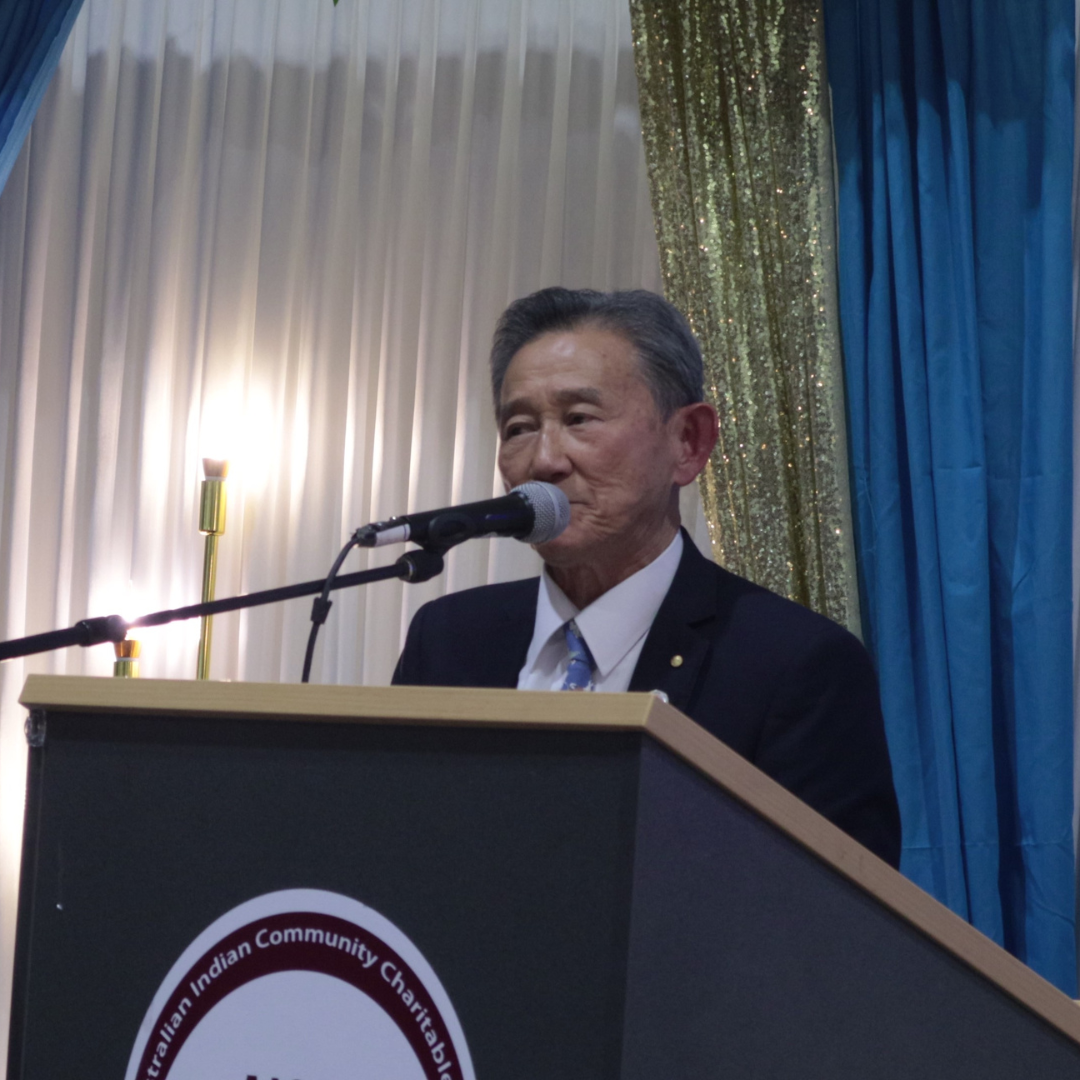 Man speaking at a podium with a microphone in front of blue and white curtains