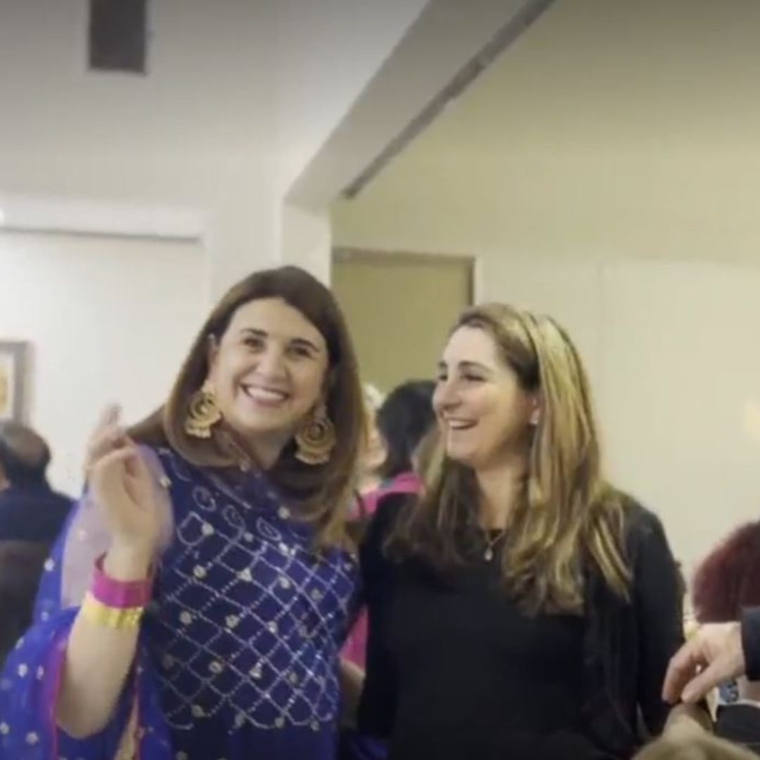 Two smiling women in a room, one waving and wearing a purple patterned top