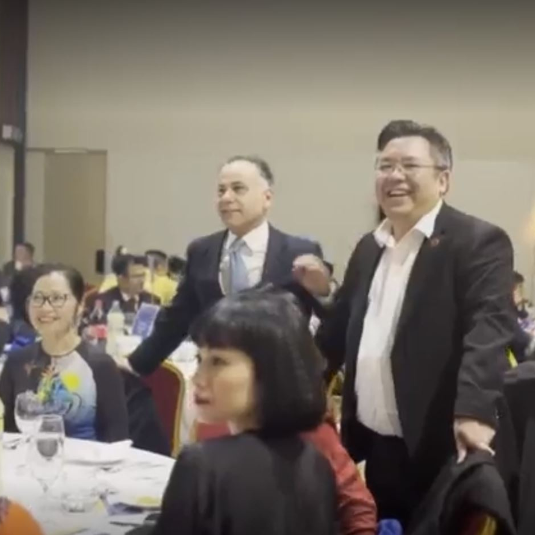 Smiling men in suits stand at a banquet table in a crowded indoor event hall.