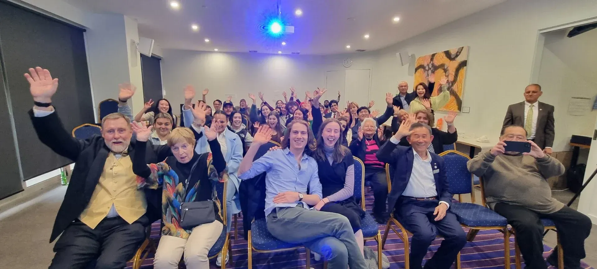 Audience in a conference room cheering and waving, with blue lights and a presenter at the front.