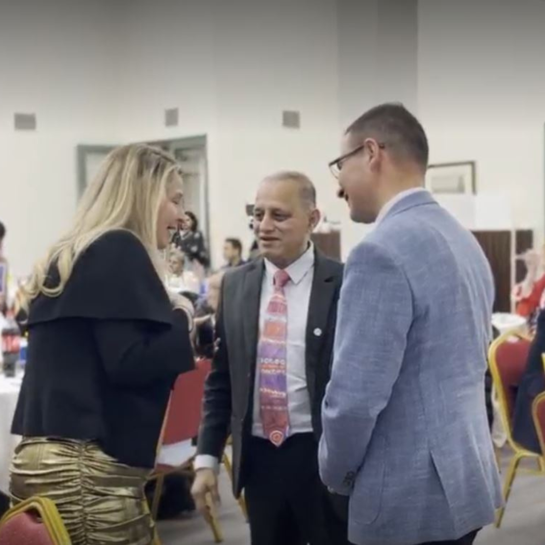 Three people talking in a bright banquet hall, with one woman gesturing and two men listening.
