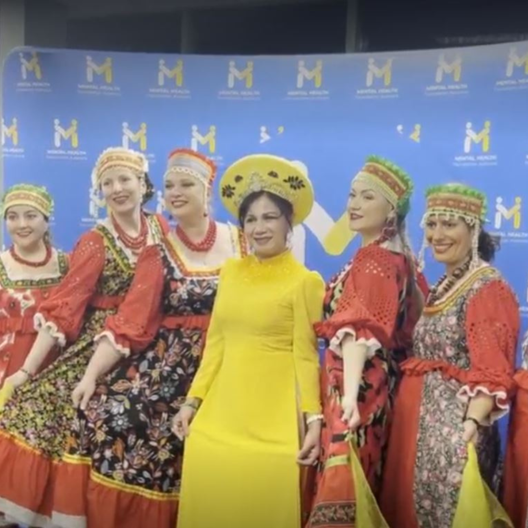 Group of performers in colorful folk costumes posing on stage before a blue backdrop with repeated logos