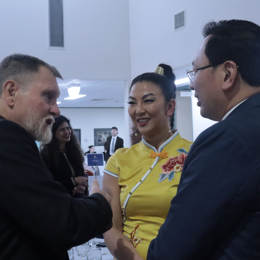 Three people conversing indoors, with a woman in a yellow dress smiling between two men.