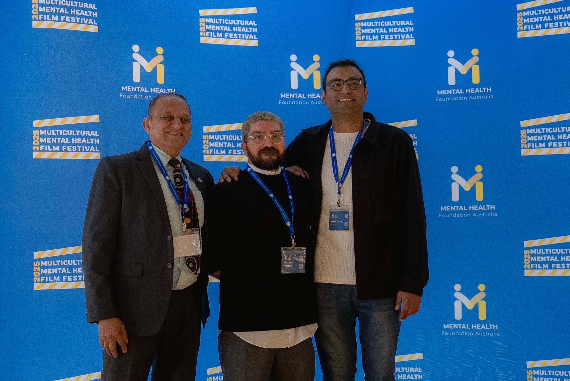 Three men standing in front of a blue event backdrop with logos, posing together.
