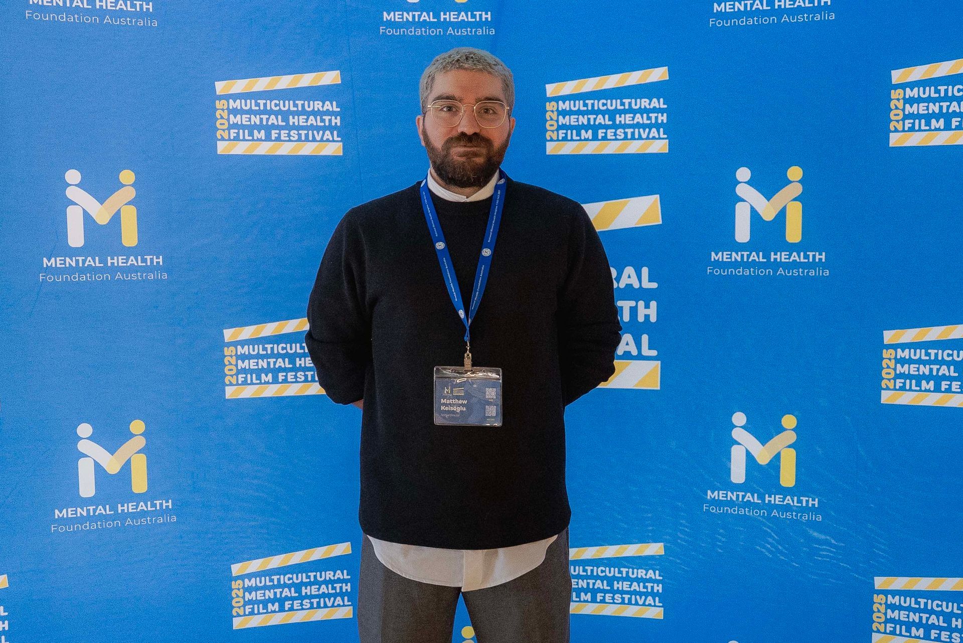 Man standing on stage before blue event backdrop, wearing black shirt and lanyard badge.