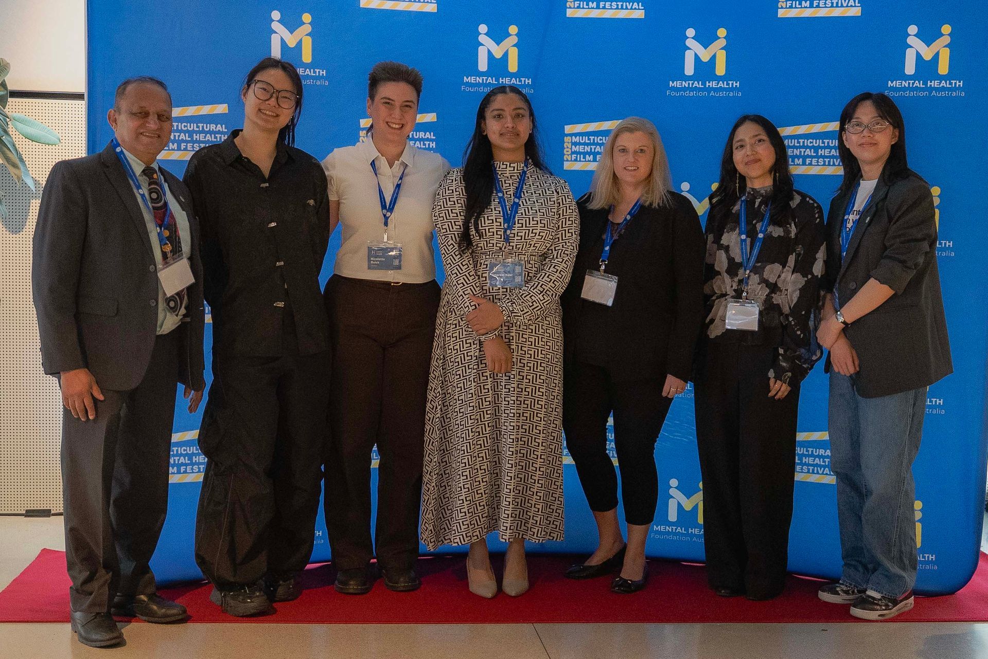 Seven people stand on a red carpet in front of a blue backdrop with repeated white “M” logos.