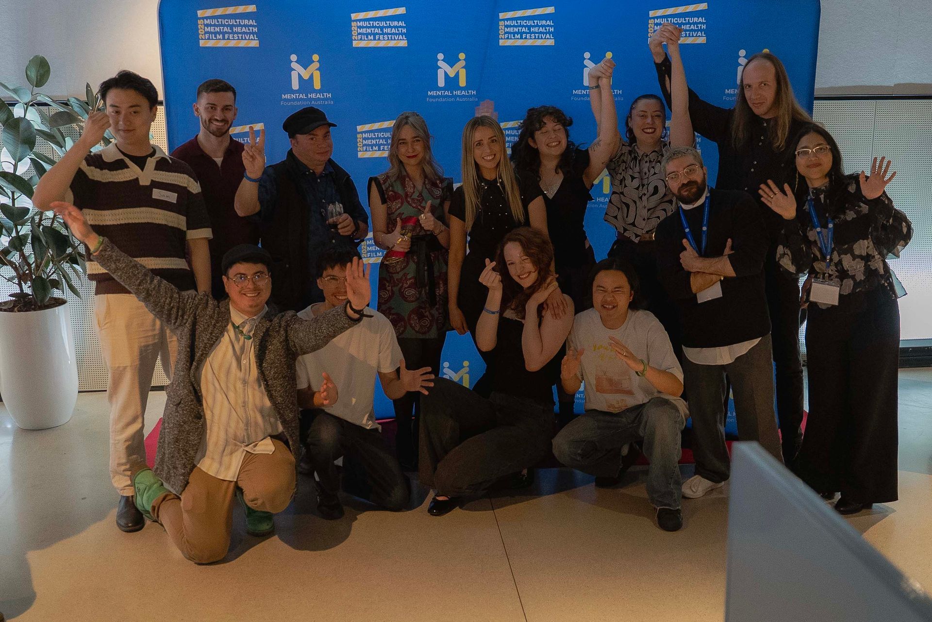 Group of people posing with a blue backdrop, smiling and waving at an indoor event.