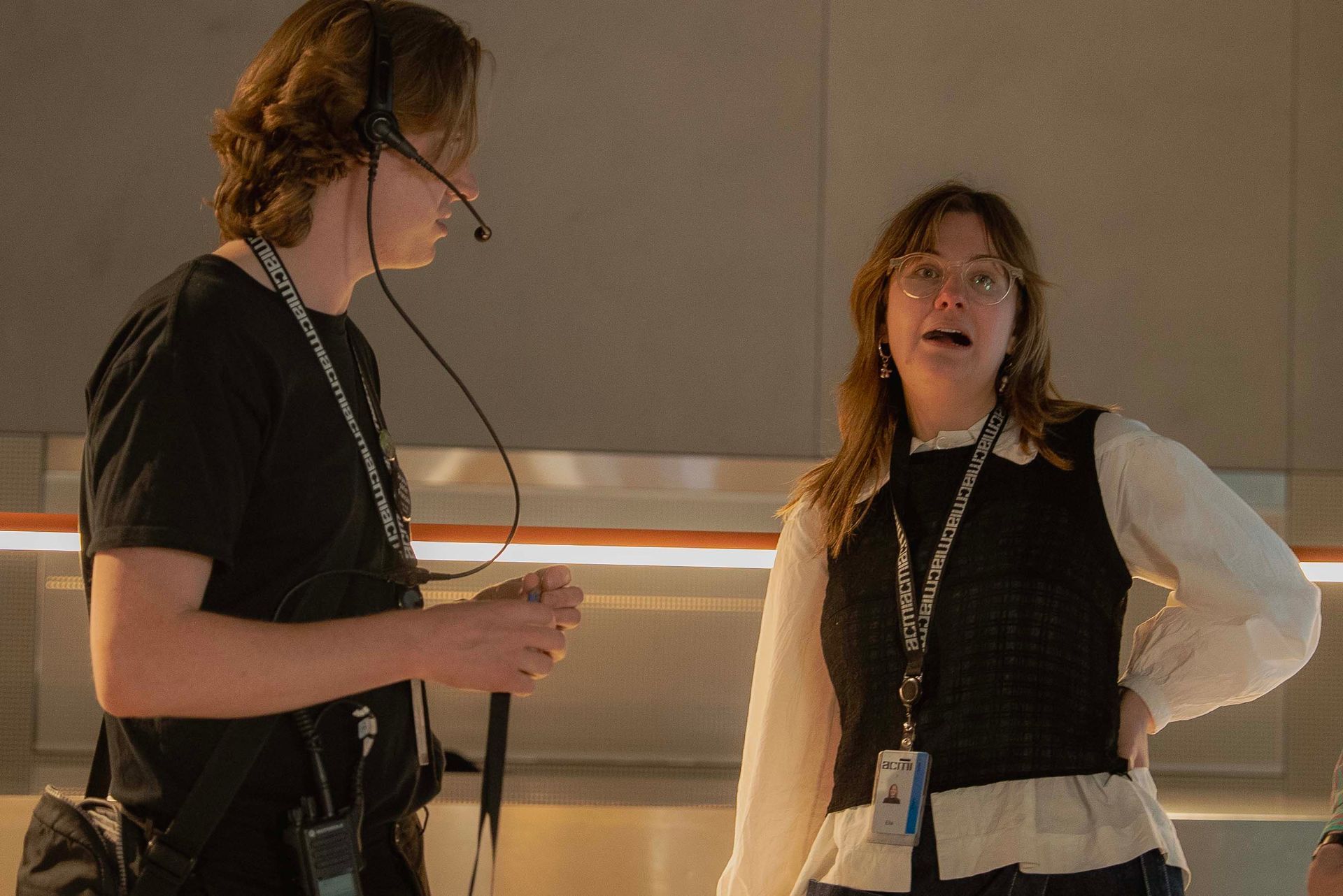 Two people in dark vests with lanyards talking in a warm-lit indoor room