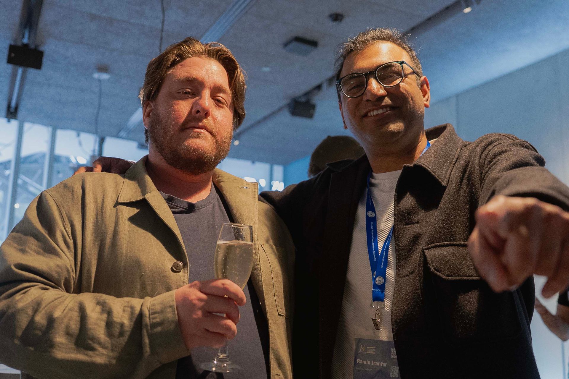 Two people posing indoors, one holding a drink and the other pointing at the camera.