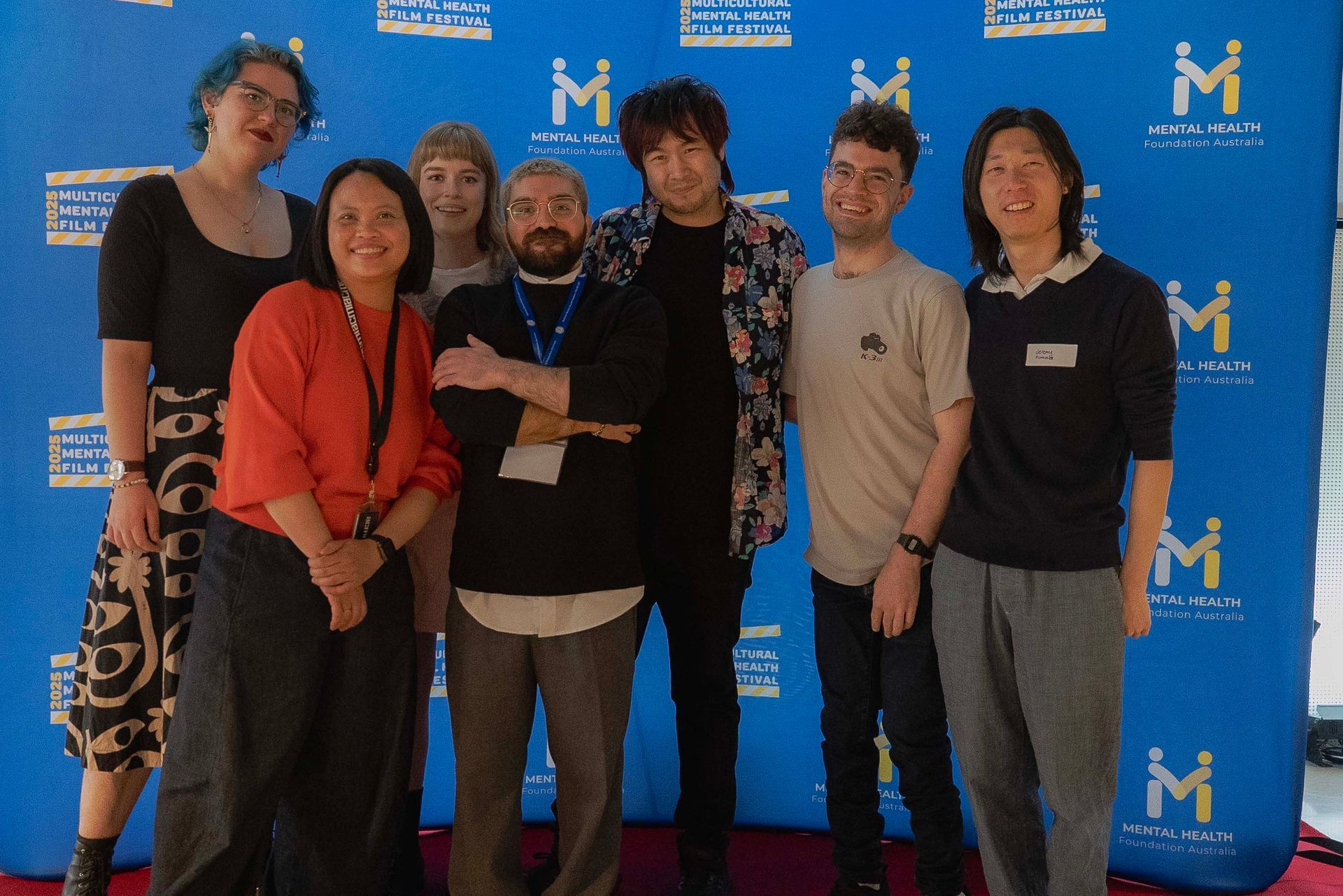 Group of six people standing and smiling in front of a blue event backdrop with logos