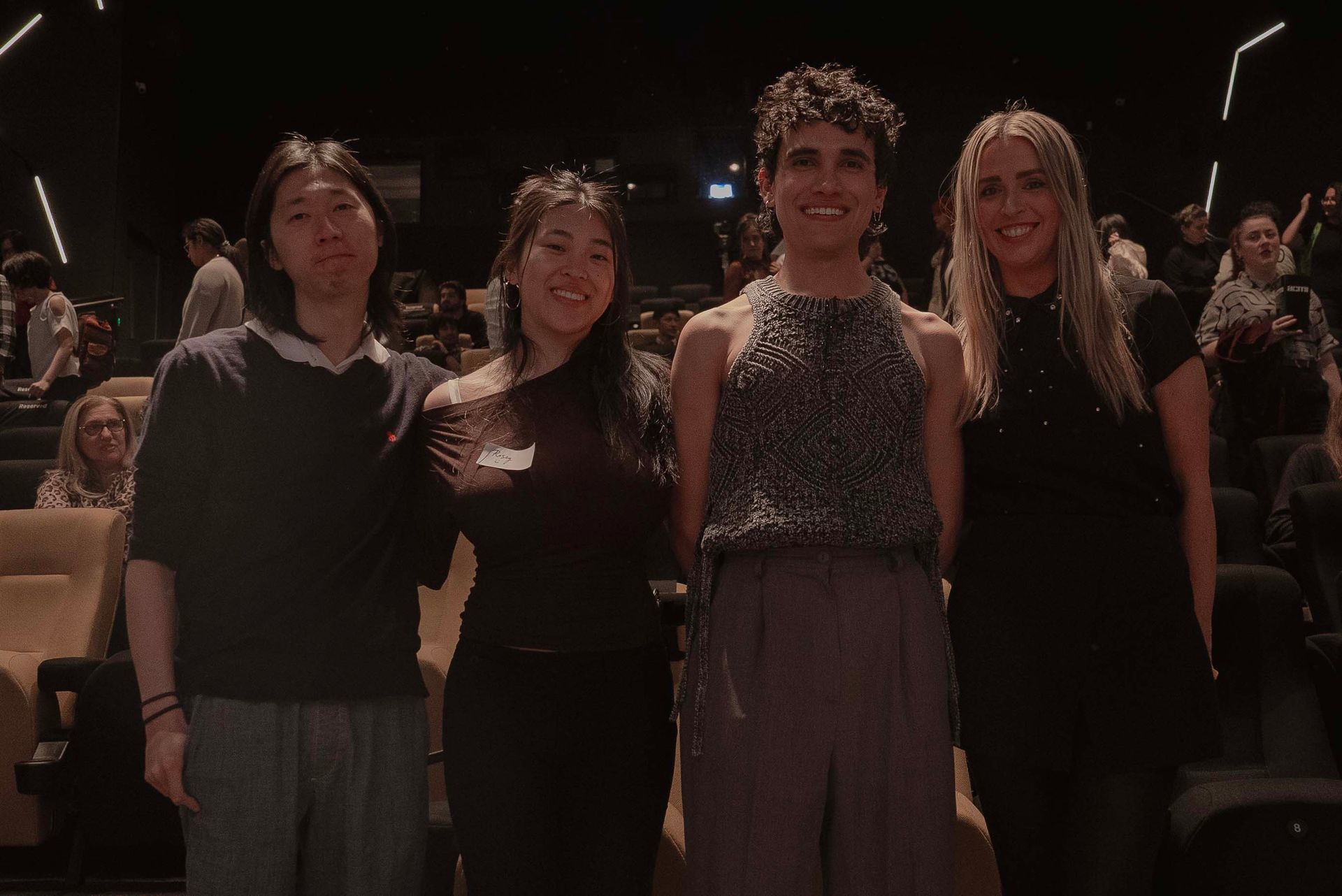 Four people standing together in a dim auditorium, posing and smiling at the camera.