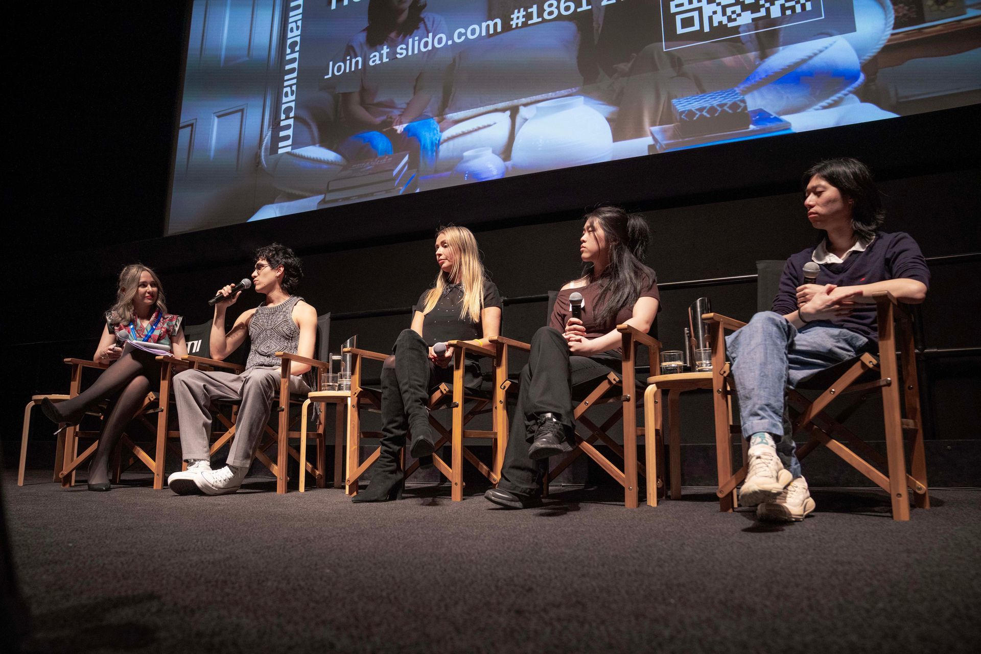 Five speakers seated on stage in director’s chairs during a panel discussion.
