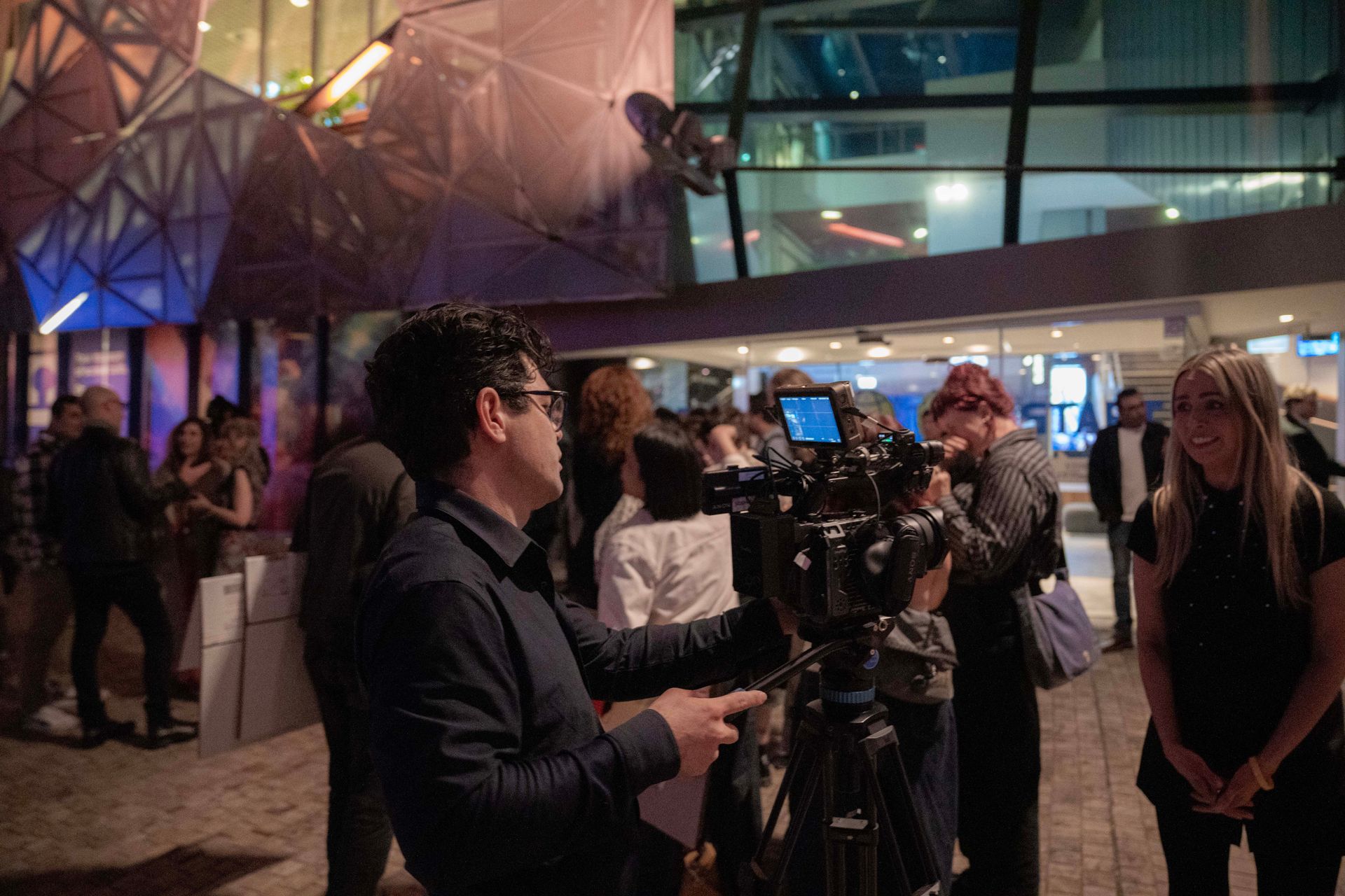 People gathered with cameras in a dimly lit modern lobby with geometric glass walls