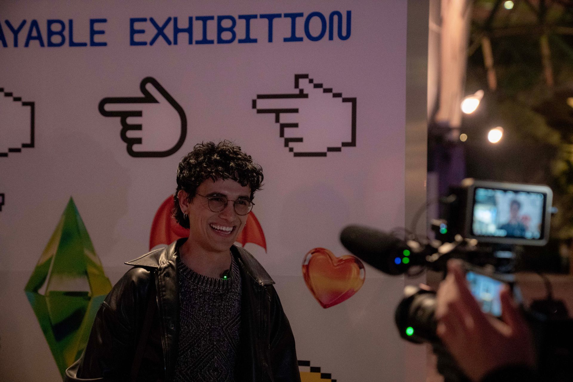 Person posing at an exhibition booth with large hand icons and a heart graphic while being filmed on a phone