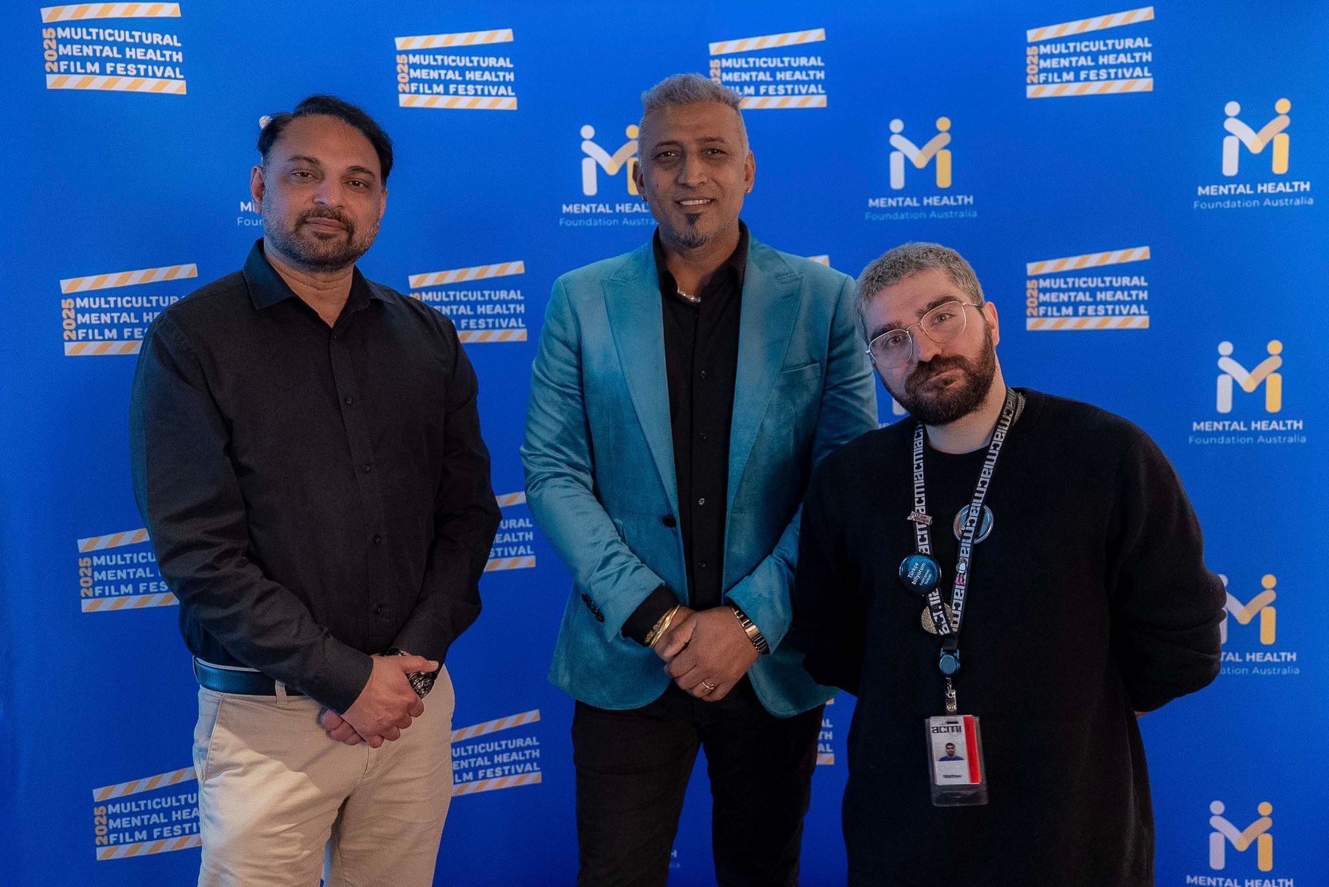 Three people posing in front of a blue event backdrop with logos, two in black and one in a blue blazer.