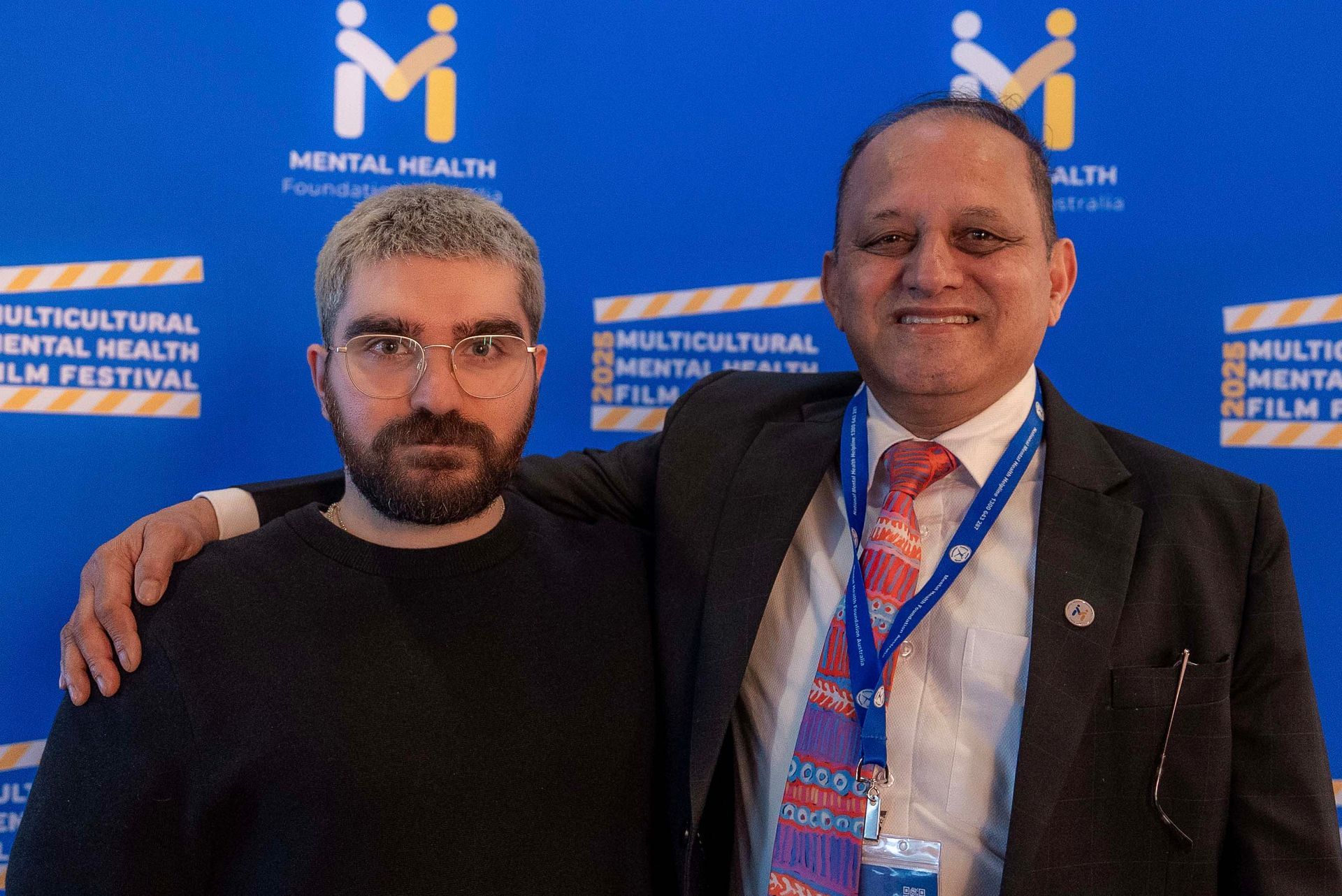 Two men posing at a Multicultural Mental Health conference backdrop, one in glasses and a black shirt, one in a suit and tie.