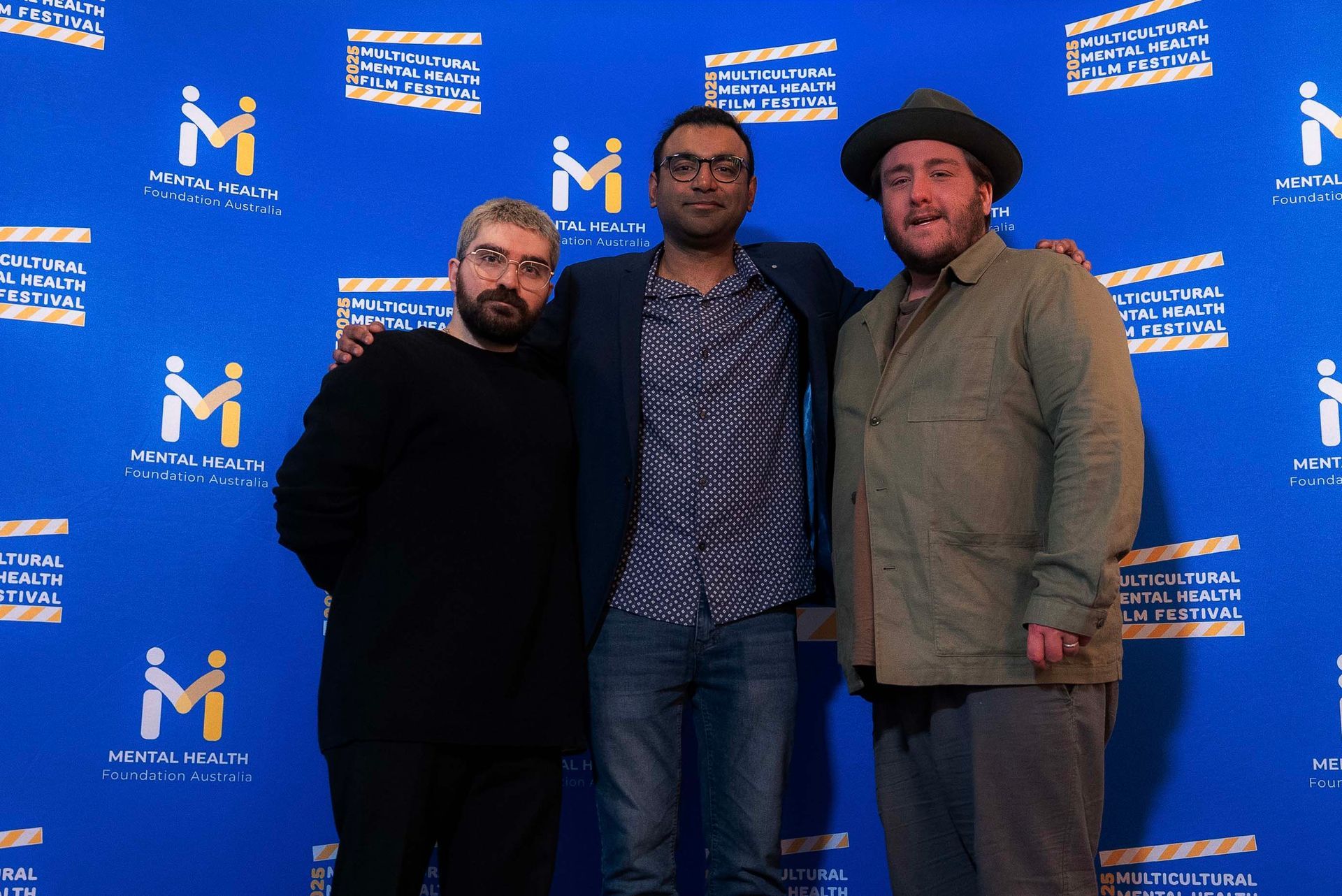 Three people posing at a blue event backdrop with repeated white and yellow logos.