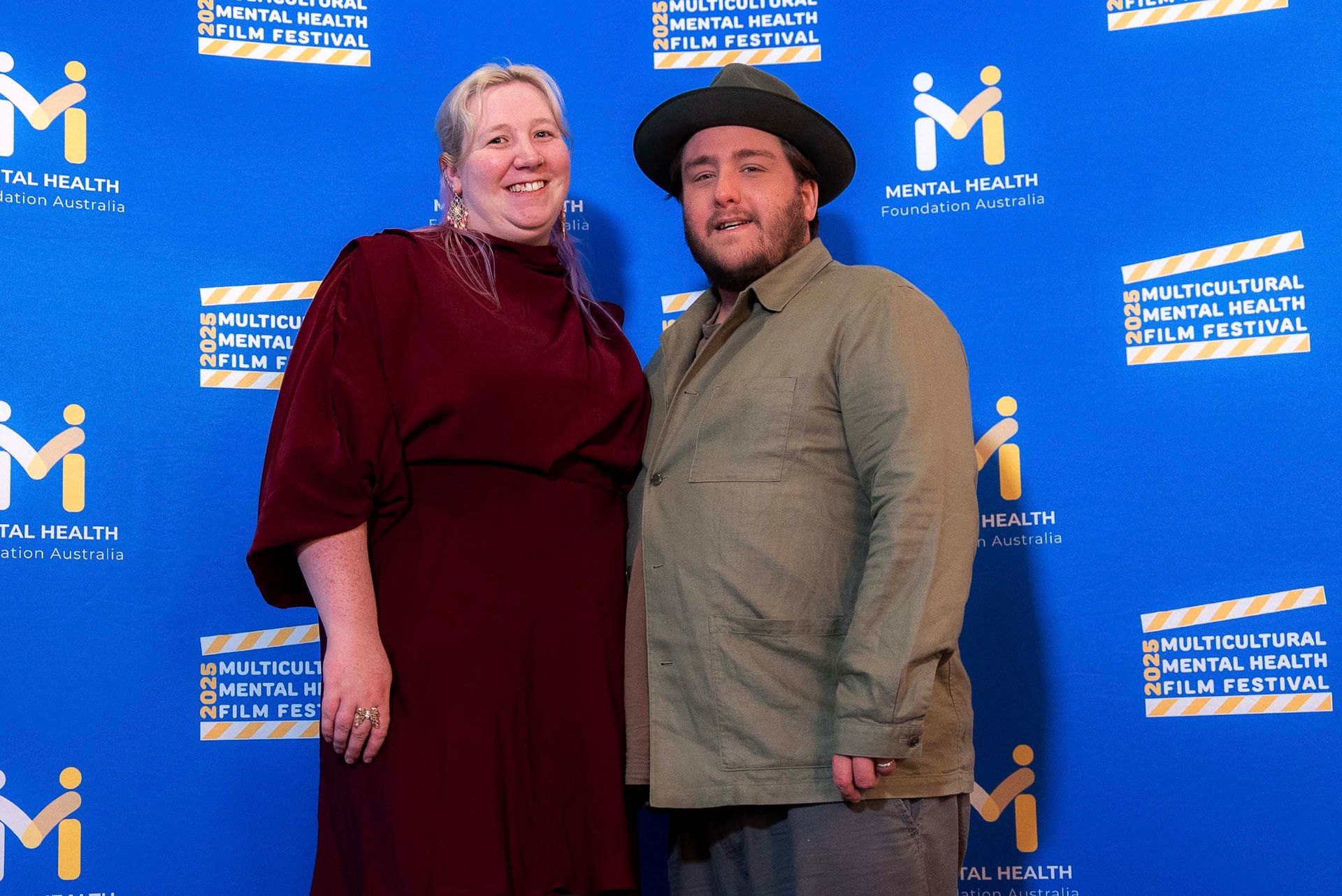 Two people posing on a blue event backdrop, one in a burgundy dress and one in a gray jacket and black hat.