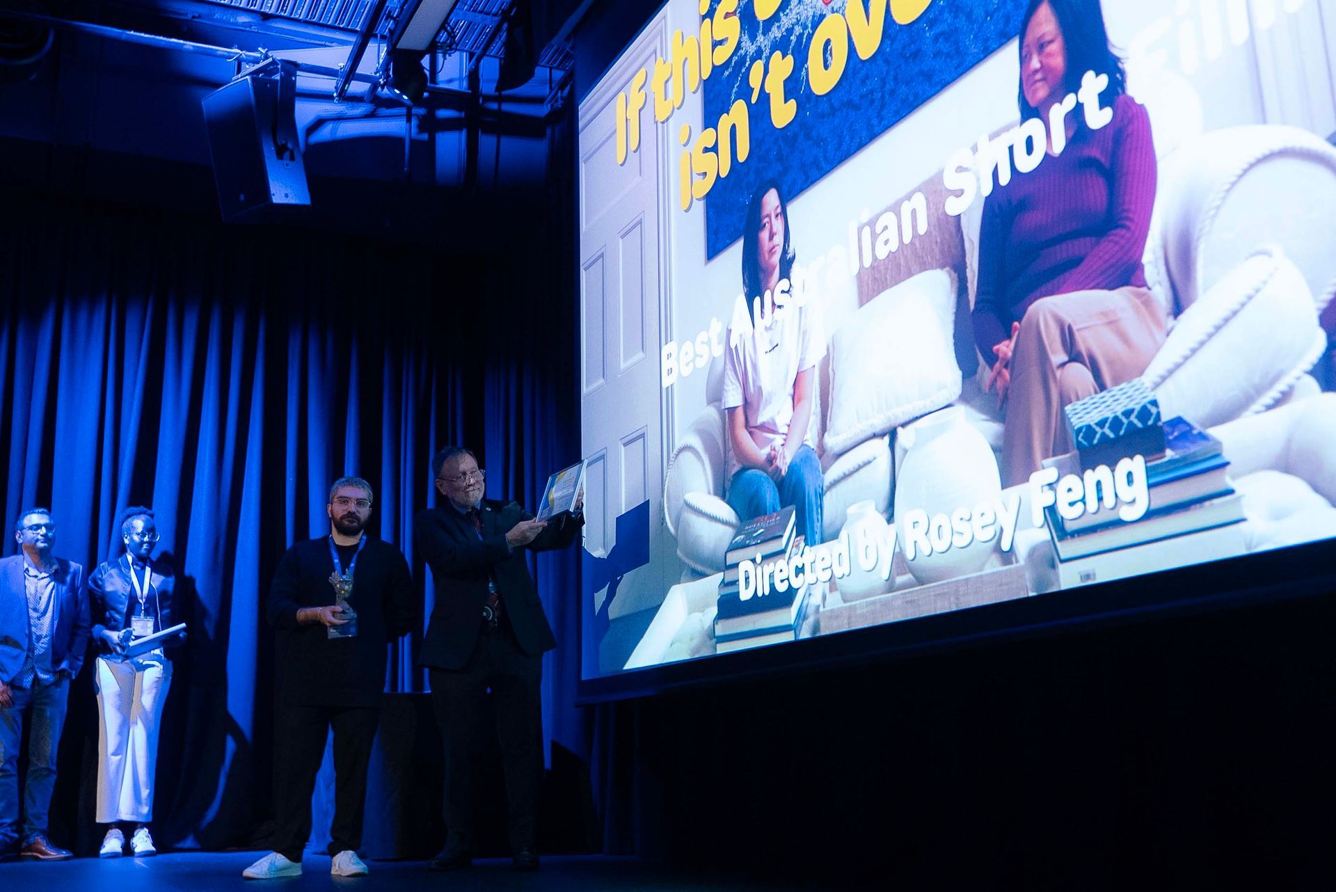 People on a blue-lit stage beside a large presentation screen showing a seated speaker and bold text.