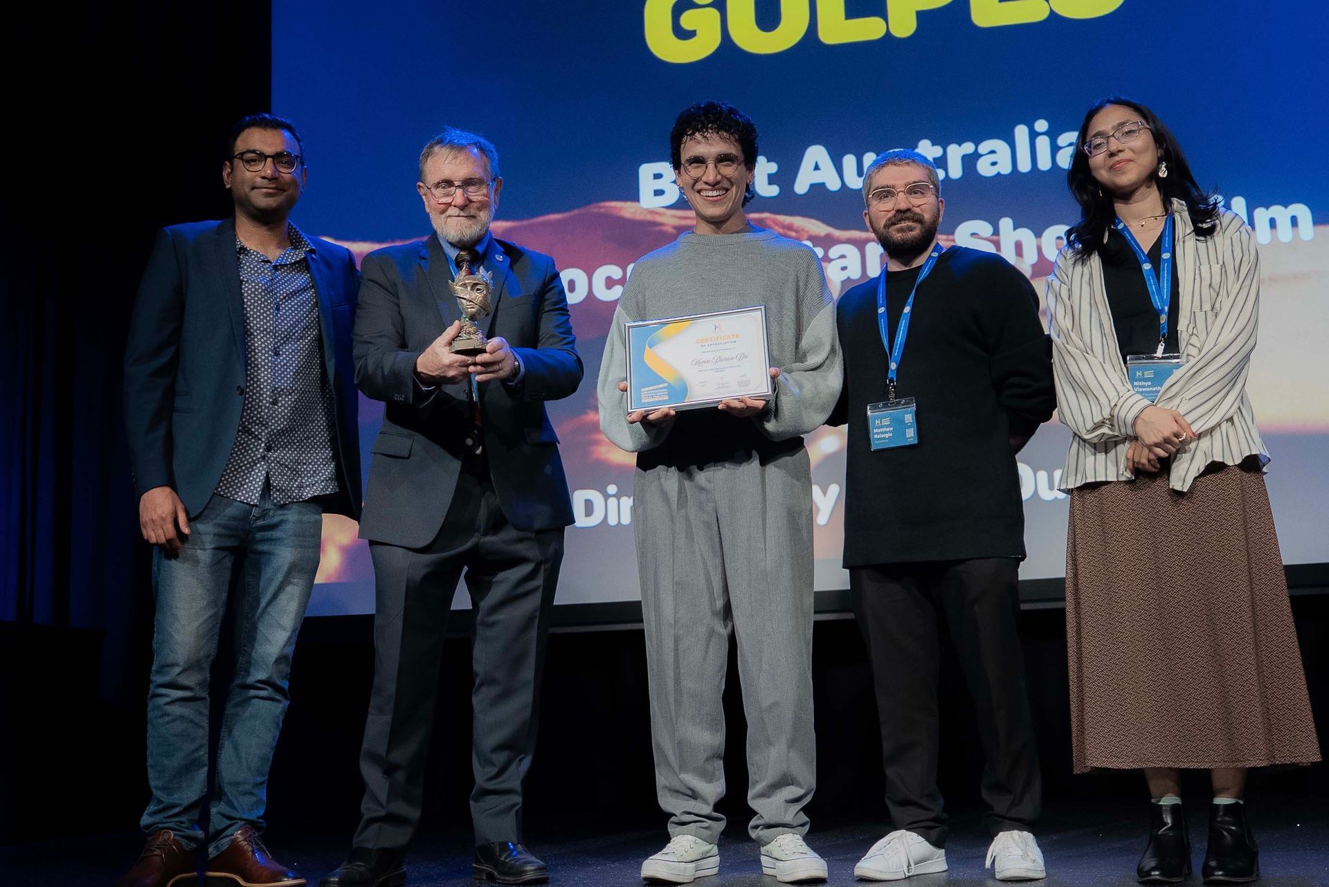 Five people on stage at an awards ceremony, one holding a certificate and another a trophy, with a GULP backdrop.