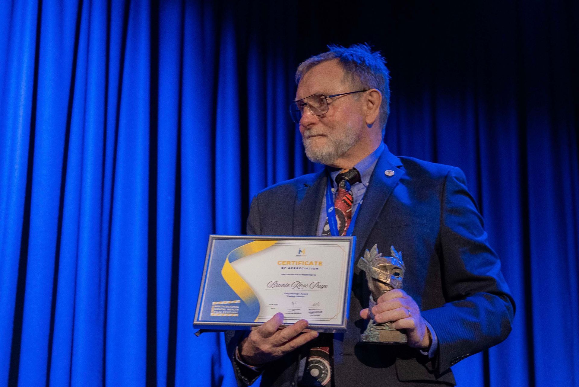 Man holding a framed certificate and trophy on stage in front of blue curtains
