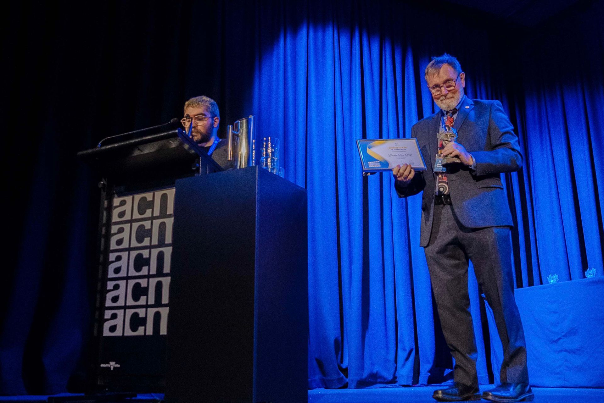 Two men on stage at a podium, one speaking and one holding notes, under blue curtains.